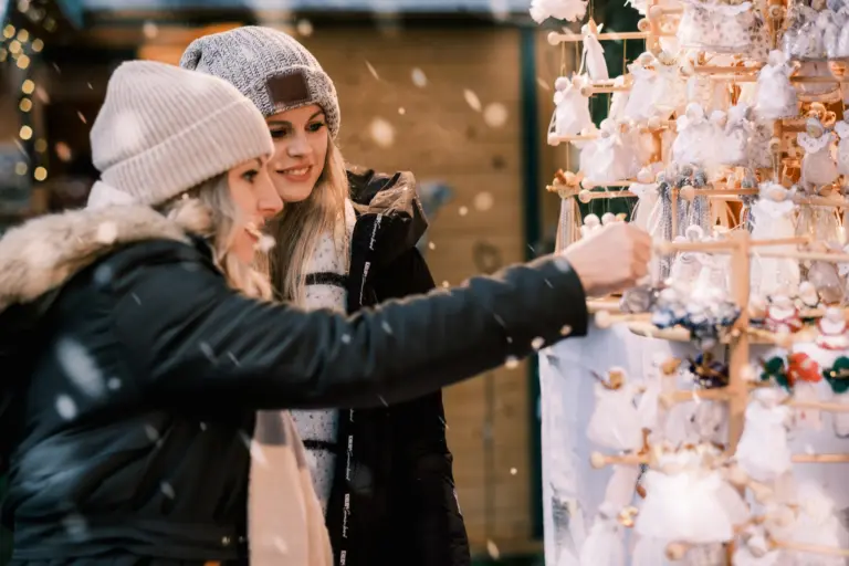 Zwei Frauen in Winterbekleidung stöbern bei Schneefall an einem Weihnachtsmarktstand mit handgemachten Engel-Anhängern.