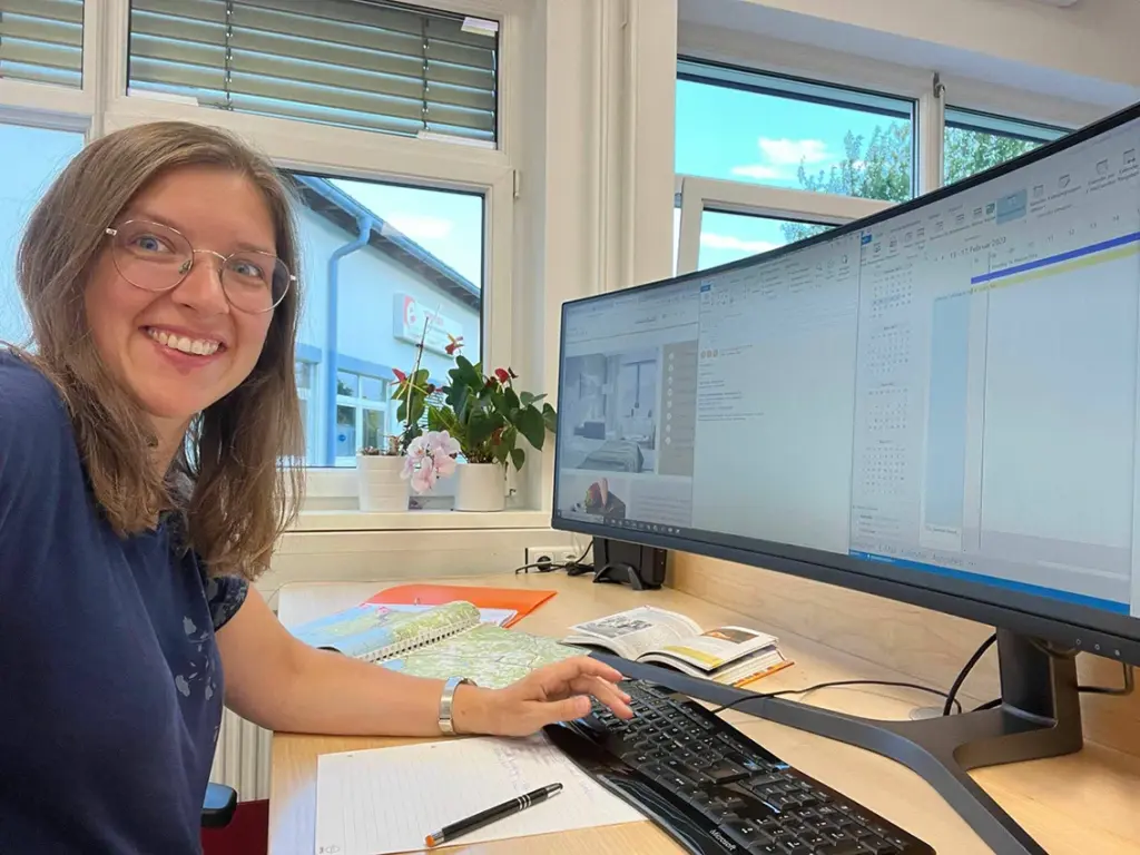 Eine in die Kamera lächelnde Frau sitzt an Ihrem Computer, Notizblock und Atlas griffbereit.