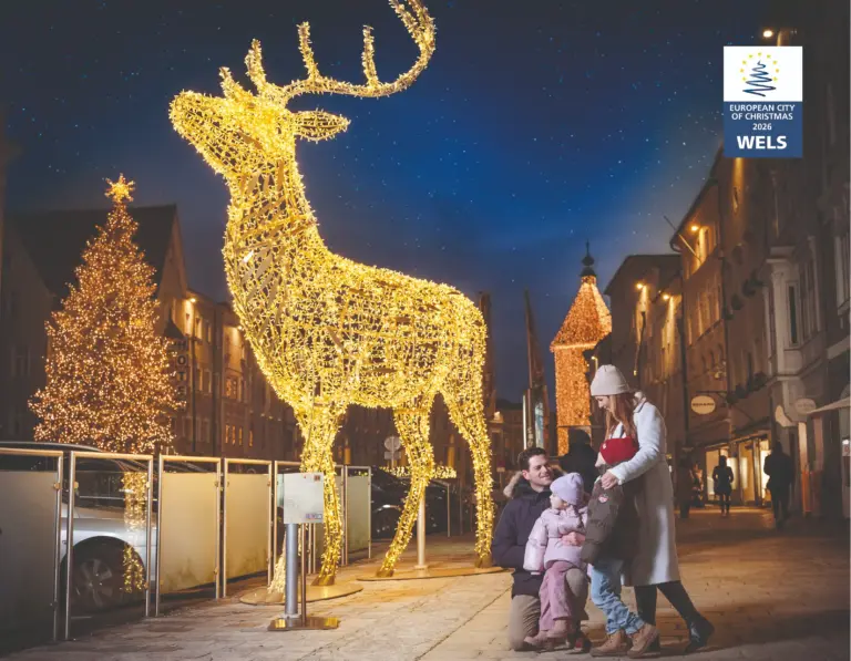 Eine große Renntier-Lichtskulptur befindet sich mittig und eine Familie die sie sich anschaut im Hintergrund sind Christbaum und festlich beleuchtete Häuser.